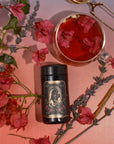 A cylindrical tin from The Lovers Box, adorned with illustrated roses and golden accents, is encircled by vibrant red and pink bougainvillea flowers, aromatic lavender sprigs, and cinnamon sticks. Close by, a glass cup of reddish tea garnished with bougainvillea petals rests on a gradient pink surface.