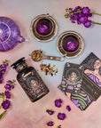 An overhead view of a vibrant tea setup featuring a purple teapot and two cups with decorative purple flowers, a jar of tea leaves, a metal tea strainer, a card with an illustration of The Moon, and a booklet titled "Magic Hour." Bright flowers embellish the scene.