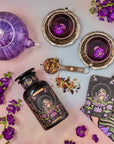 A colorful flat lay features a purple teapot and two teacups filled with herbal tea, surrounded by flowering purple plants. A jar of organic "The Moon - Tea for Nurturing Graceful Phases Case Packs" from Magic Hour, a tarot card labeled "The Moon," and a book titled "Magic Hour" complete the whimsical scene.