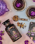 A colorful flat lay features a purple teapot and two teacups filled with herbal tea, surrounded by flowering purple plants. A jar of organic "The Moon - Tea for Nurturing Graceful Phases Case Packs" from Magic Hour, a tarot card labeled "The Moon," and a book titled "Magic Hour" complete the whimsical scene.