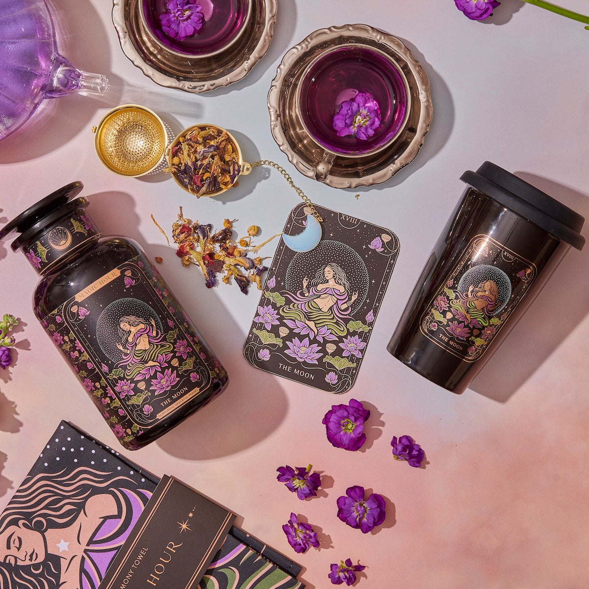 A flat lay of a mystical-themed setup features the Tea and Tarot to Go Gift Set from Magic Hour, including a black bottle, travel mug, tea infuser, tea cups filled with a purple-hued unique tea blend, flowers, a card showcasing "The Moon" tarot archetype design, and a cosmic-themed book. The items are arranged on a pink gradient background.