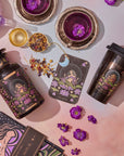 A flat lay of a mystical-themed setup features the Tea and Tarot to Go Gift Set from Magic Hour, including a black bottle, travel mug, tea infuser, tea cups filled with a purple-hued unique tea blend, flowers, a card showcasing "The Moon" tarot archetype design, and a cosmic-themed book. The items are arranged on a pink gradient background.