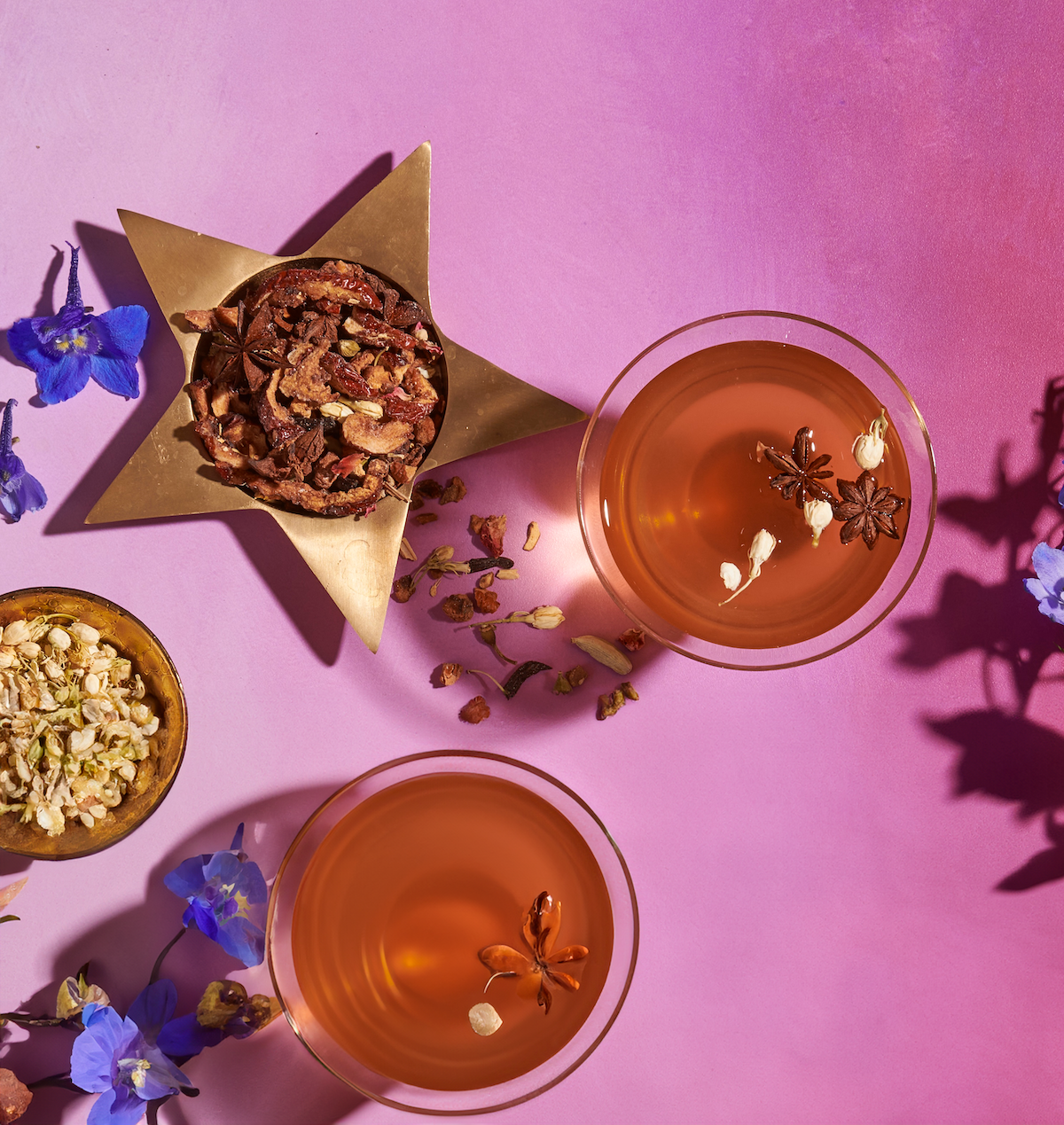 Two glasses of orange-colored "The Star: Vanilla-Ginger Beauty Potion with Jasmine & Shatavari" from Magic Hour, garnished with star anise flowers and other dried botanicals, placed on a pink background. A gold star-shaped container filled with mixed dried botanicals and a small bowl of herbs are nearby. Blue and purple flowers are scattered around.