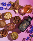 A flat lay of The Star Box items, including assorted spices and ingredients arranged in small bowls on a pink surface. It features an artisan tea strainer, an artistically designed bottle, a glass of the Star Vanilla Ginger Beauty Potion, a star-shaped bowl of dried herbs, and scattered blue flowers.