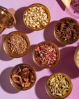 An arrangement of six bowls filled with various dried ingredients on a pink surface. The items include seeds, spices, and dried flowers, perfect for crafting your own loose leaf tea using The Star: Vanilla-Ginger Beauty Potion with Jasmine & Shatavari from Magic Hour. A brass mortar and pestle, along with a triangular cloth bag, are also present in the composition.