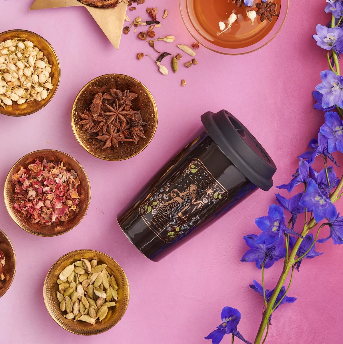 A black travel mug featuring decorative designs rests on a pink surface, surrounded by various small bowls filled with different spices and dried herbs. To the right, clusters of vibrant blue flowers perfect the scene for an enchanting Magic Hour Tea and Tarot to Go Gift Set experience.