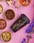 A travel mug with a celestial design is on a pink surface surrounded by bowls containing various dried spices, including star anise, cardamom, and rose petals. Nearby, there's a cup of organic tea and a few pastries, while vibrant blue flowers also adorn the scene.