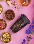 A black travel mug featuring decorative designs rests on a pink surface, surrounded by various small bowls filled with different spices and dried herbs. To the right, clusters of vibrant blue flowers perfect the scene for an enchanting Magic Hour Tea and Tarot to Go Gift Set experience.