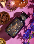 A dark bottle with a label featuring "The Star" tarot card design lays on a pink surface surrounded by various dried herbs in small bowls, a cup of Magic Hour tea, and purple blue flowers. The setup has a mystical and herbalist theme and showcases the Magic Hour product The Star: Vanilla-Ginger Beauty Potion with Jasmine & Shatavari.