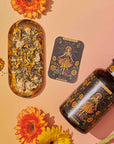 A vibrant flat lay showcases Magic Hour's tea products, "The Sun: Energizing Adaptogenic Herbal Tea with Yaupon, Moringa & Turmeric," in elegant black and gold packaging, surrounded by colorful flowers. A gold tray holds sliced fruits, a tea strainer, and a teacup. Loose Yaupon tea leaves and a golden teaspoon are artfully arranged nearby against a gradient yellow-pink background.