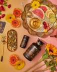 A flat lay featuring The Sun: Energizing Adaptogenic Herbal Tea with Yaupon, Moringa & Turmeric in a floral tin, a golden teapot, two black gold-detailed containers, fresh flowers, and a fruit tray on a pink and yellow background.