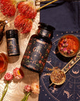 An ornate setup featuring The Wheel of Fortune Box with a large black jar and a smaller container adorned with intricate designs, surrounded by various herbal ingredients, flowers, and a golden tea set. There's a celestial-themed mat underneath, adding an astrological ambiance to the scene.