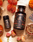 An arrangement featuring a large black jar with a floral design, a smaller matching container, a steaming glass of tea, and a teapot on a decorative surface. Surrounding the items are various vibrant flowers and herbs. Next to the jars lies The Wheel of Fortune tarot card.
