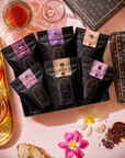The Best of Magic Hour Sampler Set with Tea in Hand Teapot, featuring an eco-luxe gift box of six tea pouches surrounded by a glass teapot, loose tea leaves, and scattered flower petals.