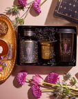 A Tea and Tarot to Go Gift Set features elegantly designed black tea containers and a gold cup, all beautifully packaged in a box adorned with stars. Accompanying the set is a teacup filled with loose leaf tea and a Ceramic Tarot Mug pouring milk. The scene is enhanced by the addition of purple and pink flowers, complemented by a piece of amethyst, creating an ideal presentation for any tea lover or tarot enthusiast.