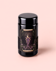 A black jar adorned with a mystical label depicting an illustrated woman encircled by flowers and stars, titled “The Empress: Lavender Currant Shatavari Cocoa Tea of Nurturing Creativity” from Magic Hour, stands out against a soft pink background. This enchanting vessel contains an organic herbal tea blend crafted to inspire wellness—a Lavender Currant Shatavari Cocoa infusion designed to nurture creativity while soothing the senses and delighting the palate.