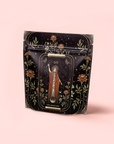 A decorative black bag with a tarot-inspired "The Magician" design, golden accents, and flowers on soft pink, perfect for storing The Magician - Elderberry-Ginger-Vanilla Herbal Tea or your favorite wellness blend.