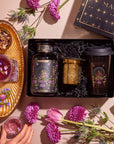 The Tea and Tarot to Go Gift Set embodies mystical elegance, with a selection including tea canisters, a travel mug, and floral design elements. A gold tray holds a cup of purple tea, surrounded by decor inspired by tarot archetypes. Amidst this enchanting setting infused with magic, a hand reaches for the cup.