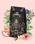 A decorative tea package for "The Moon - Tea for Nurturing Graceful Phases," adorned with elegant floral and lotus designs, is surrounded by vibrant flowers and fruit slices on a pale pink background. The packaging features a serene figure seated among lotuses, capturing the soothing essence of chamomile tea.