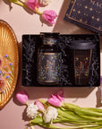 Introducing the captivating Tea and Tarot to Go Gift Set, which includes a beautiful starry jar and a Ceramic Tarot Mug arranged in an elegant black box. It is charmingly complemented by pink tulips, white flowers, an amethyst crystal, a golden tray adorned with gems, and a whimsical book about magic—ideal for those who resonate with the tarot archetype.