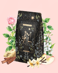 A decorative tea package featuring "The Star: Vanilla-Ginger Beauty Potion with Jasmine & Shatavari" is adorned with a star tarot card design and surrounded by flowers, apple slices, vanilla beans, and cinnamon sticks on a soft pink background—perfect for highlighting this organic herbal blend.