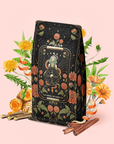 A tarot-inspired package of "The Wheel of Fortune: Ginseng Spice Tea for Adapting to Change" sits amid orange flowers, dandelions, cinnamon sticks, and roots on a soft pink background—this detox adaptogenic blend brings a unique twist to tradition.