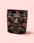 A black resealable pouch with ornate red flowers and gold accents displays a tarot-inspired Wheel of Fortune and Egyptian sphinx, holding The Wheel of Fortune: Ginseng Spice Tea for Adapting to Change, set against a light pink background.