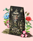A decorative package of "The World: Tea of Infinite Possibiliteas" black tea, adorned with tarot-inspired artwork and surrounded by flowers, loose tea leaves, and rose petals on a soft pink background. Infused with notes of Blue Lotus, this elegant design invites you to savor an enchanting and magical brew.