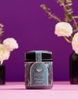 A Ceremonial Bath Salt Soaks - Violet Glass Jar sits on a pink surface with a purple background, flanked by two black vases with white flowers and greenery—ideal for enhancing your self-care ritual.