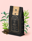 A black bag of "Ti Quan Yin Oolong: Tea of Patient Compassion" rests before green leaves and white tea flowers, with loose oolong scattered on a pink background. The package shows a gold hourglass and celestial symbols, celebrating wellness benefits.