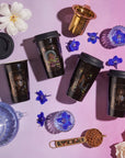 Five black travel mugs with artistic designs are placed on a pink surface, surrounded by blue flowers, a white flower, a clear glass cup, a blue teapot, and a brass tea infuser. A spoon filled with Magic Hour’s The Queen of Cups - Tarot To Go! completes the enchanting scene.