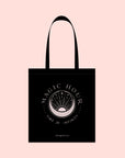 The Magic Hour Tote Bag features a black cotton design with white illustrations and text. It showcases a stylized hourglass at the center, embellished with celestial motifs in metallic gold. The words "Magic Hour" appear at the top, while "Time for Infinity" is displayed below. A soft peach background complements the mystical theme, adding warmth.