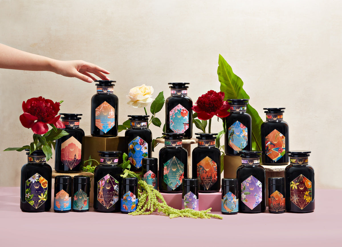 A collection of black jars adorned with colorful labels is arranged on a pink surface against a light background. The jars vary in size and are surrounded by vibrant red and white flowers, with a hand reaching toward one of the jars. Green foliage accents the scene.