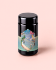 A glossy black jar labeled “Tulum Horchata Chai,” adorned with a vibrant geometric design, botanical illustrations, and gold lettering, set against a pale pink background—an elegant vessel for an adaptogenic wellness potion infused with ashwagandha and yerba mate.