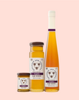 Three Savannah Bee Co. Honey - Tupelo jars from the Apalachicola River Basin sit on a light pink background. The smallest jar is open, the medium jar is closed, and the tallest bottle has a corked long neck. Each label showcases an intricate honey bee illustration.