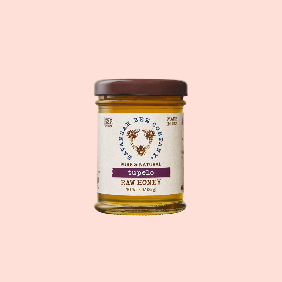 A jar of Savannah Bee Co. Honey - Tupelo, harvested from the Altamaha River Basin, features a brown lid against a light pink background. The cream-colored label displays black and purple text with a bee logo and floral design.