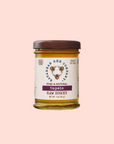 A jar of Savannah Bee Co. Honey - Tupelo, harvested from the Altamaha River Basin, features a brown lid against a light pink background. The cream-colored label displays black and purple text with a bee logo and floral design.