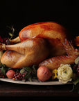 A whole roasted turkey sits on a platter with flowers, greenery, berries, and peaches, its flavor enriched by Lapsang Souchong Turkey Brine, set against a dark background.