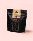 A black and gold stand-up pouch labeled "Bohemian Breakfast Vanilla Puerh Black Tea for Digestion & Energy" by Magic Hour features a celestial design, highlighting this organic tea as a dreamy coffee alternative against a pale pink backdrop.