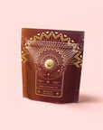 A maroon pouch with gold geometric and sunburst designs, labeled "Queen of the South Herbal Adaptogen Tea," is featured on a pink background—an ideal addition to the Herbal Tea Sampler Tea Box: Caffeine-Free Teas for Sleep, Digestion & Immunity.