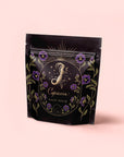 A black resealable bag with gold and purple floral designs, labeled "Capricorn: Maple Oolong Tea" and "Magic Hour," features a crescent moon, stars, and an ornate letter "J"—crafted with metabolism-boosting Se Chung Oolong on a pale pink background.