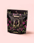 A decorative black pouch with floral designs shows two nude figures embracing under an arch, surrounded by purple flowers and leaves. The label reads "The Lovers Hibiscus-Cinnamon-Vanilla Love Potion" with organic hibiscus and balancing herbs.