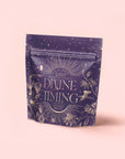 A purple resealable tea pouch labeled "Divine Timing Rest" displays gold celestial and botanical designs on a pale pink background, indicating a relaxing nighttime herbal adaptogen tea blend to support restful sleep.