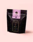 A black resealable pouch with gold star patterns labeled "Magic Hour" and "Time + Infinity," featuring a purple label that reads "Antioxidant-rich Almond Matcha Green Tea for Joy." The bag is set against a pale pink background.