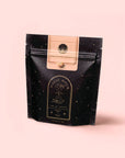 A black and gold resealable pouch labeled "Magic Hour" with celestial designs and "Goddess Green Tea" text sits on a pale pink background. The mystical packaging hints at flavors of apricot lychee and organic lemongrass.