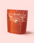 A red resealable pouch with a gold cosmic design—radiating lines, circles, and hands around a sun—on a pale pink background. The label reads: "Queen of the Harvest : Herbal Relaxation Tea. Caffeine-Free.