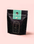 A standing black resealable pouch labeled "Magic Hour" with a celestial design and green top label showcases Mantra Mint™ Herbal Tea, featuring calming Ashwagandha. Set against a pale pink background, this blend is naturally caffeine-free.