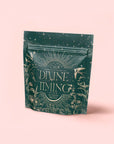 A green tea package labeled "Divine Timing Calm" features gold plant, star, and hummingbird illustrations on a pale pink background. This premium botanical wellness tea contains a cooling mint adaptogen blend for calm and emotional balance.