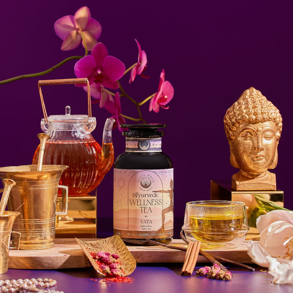 A bottle of Vata Herbal sits among a teacup, teapot, golden mortar and pestle, honey jar, cinnamon sticks, dried flowers, and a golden Buddha statue on a purple background with orchids.