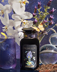 A glass of purple Vanilla Lavender Black Iced Tea with a lemon slice, a Cosmic Garden Iced Teas bottle, white orchids, lavender sprigs, a butterfly, and two glass orbs set against a starry cosmic backdrop.